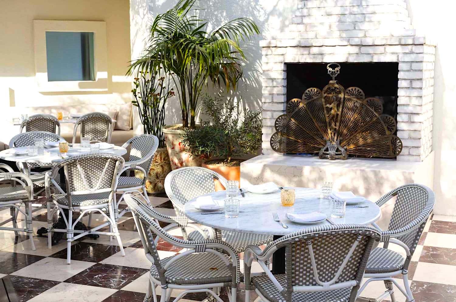 A cozy outdoor lounge with a white brick fireplace, metal screen, wicker chairs, marble tables, and a potted plant.