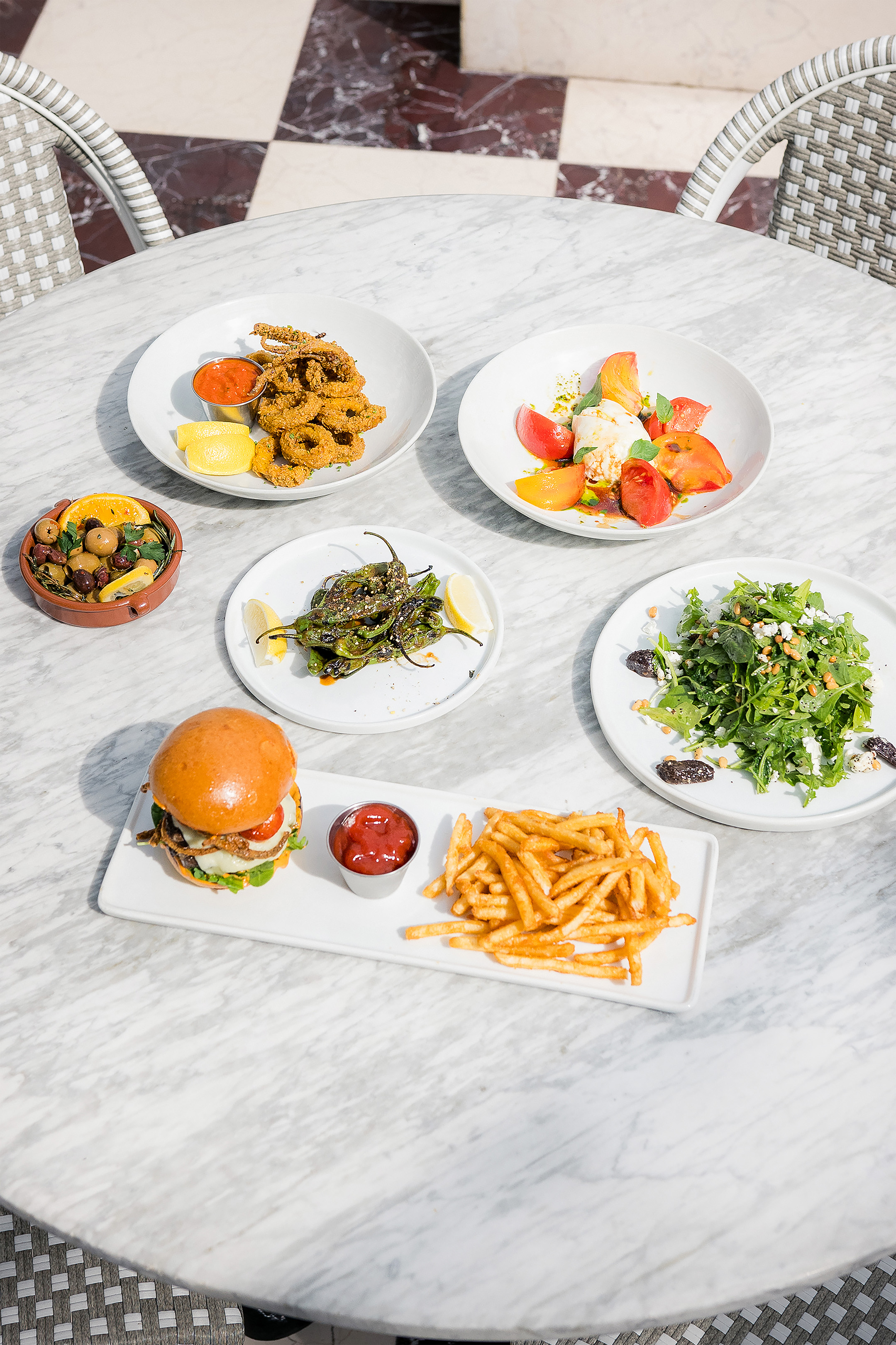 A table set with various dishes, including a burger, fries, salad, and other food items, on a marble table with wicker chairs.