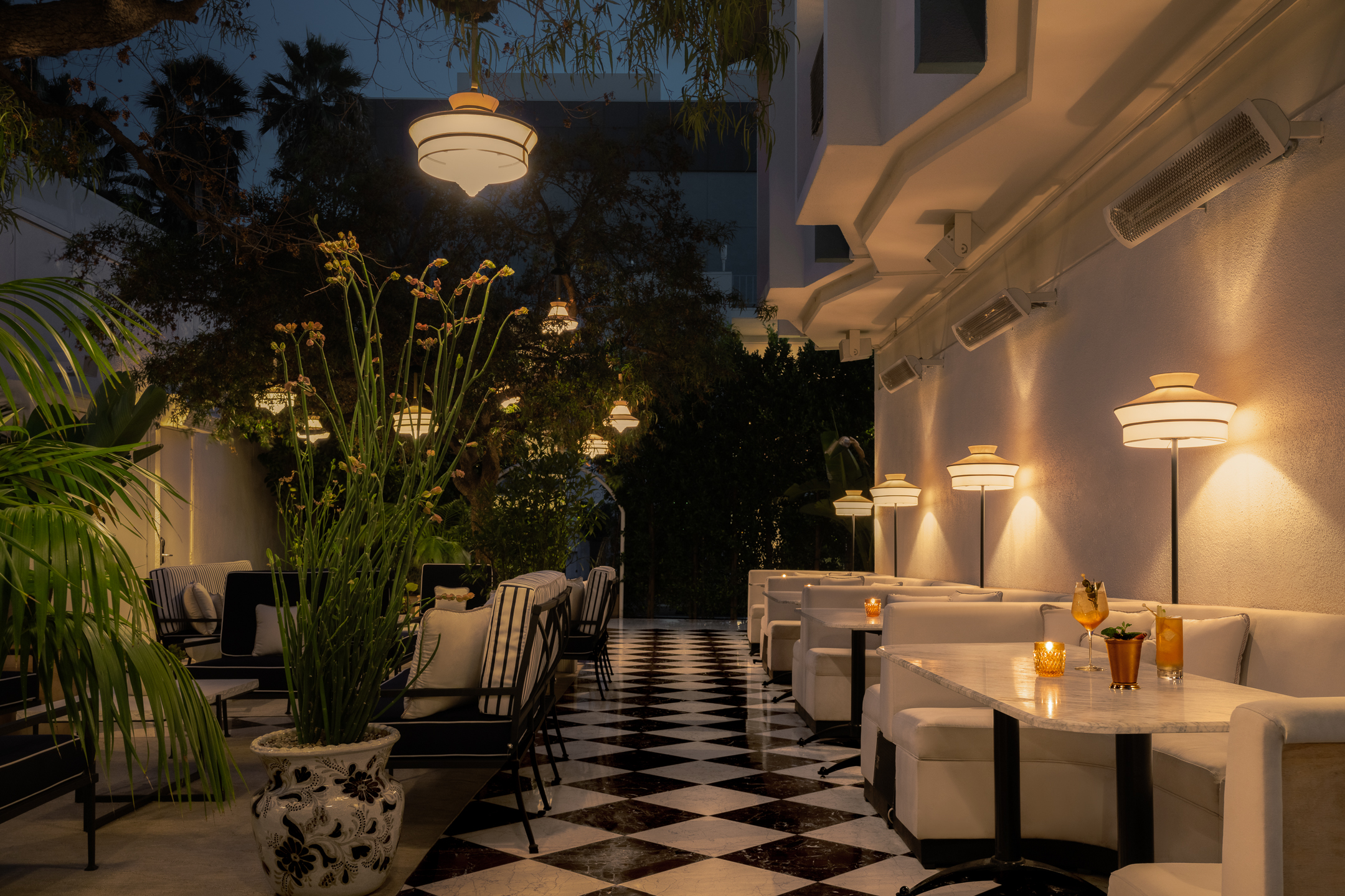 A dimly lit bar with white marble tables and white leather banquette seating, with hanging lights and potted plants.