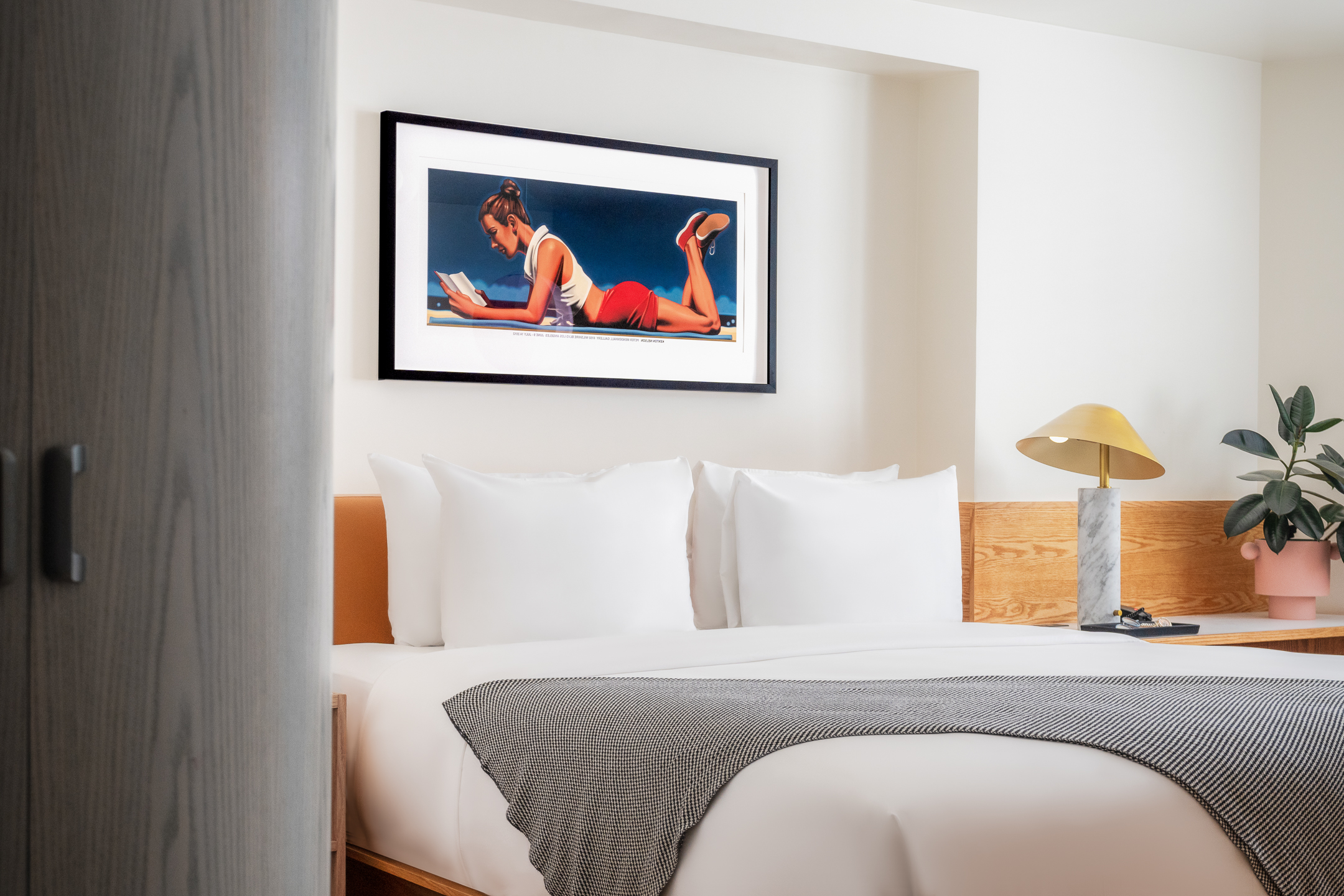 A hotel room with a bed, nightstand, lamp, plant, and artwork