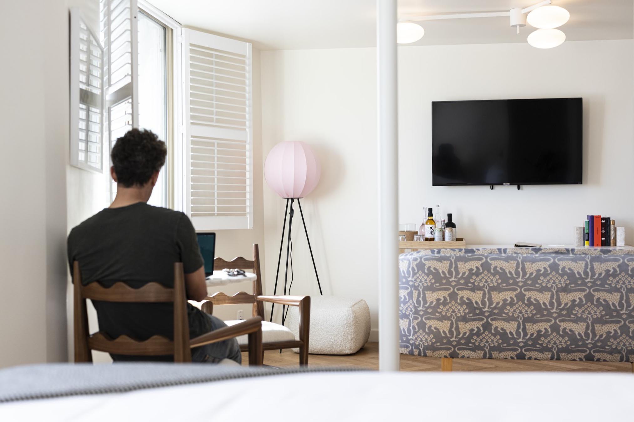 A man sits at a desk in a hotel room with a TV, lamp, and window.